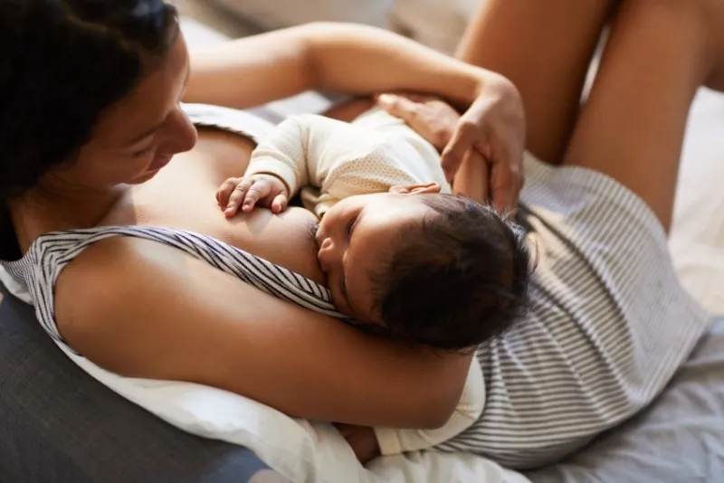close-up of serene loving young black mother in nightgown sitting on bed and breastfeeding baby in comfortable position