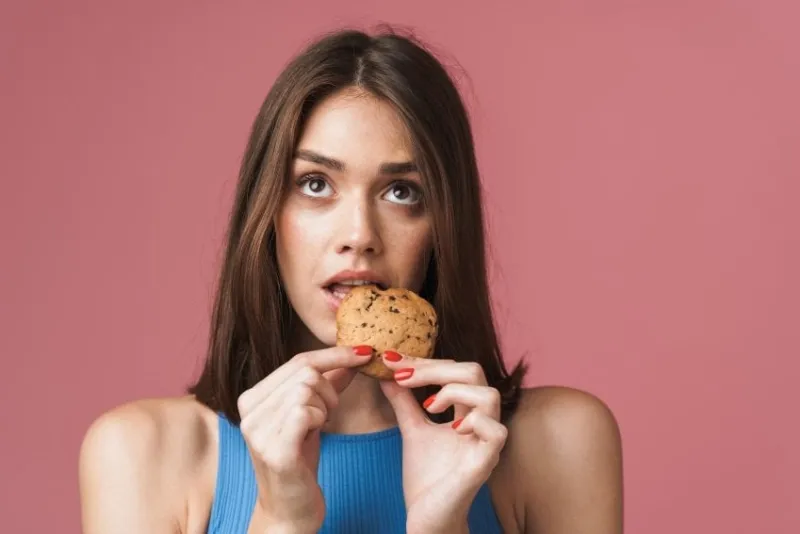 portrait of a young attractive brunette woman standing isolated over pink background, eating a chocolate chip cookie, looking up