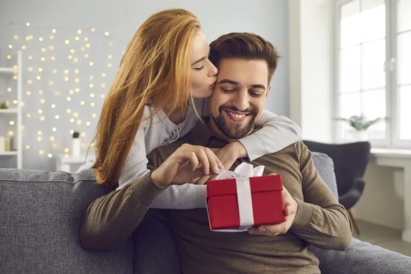 love background young redhead wife hugs and kisses her husband on the cheek giving him a gift for valentine's day smiling bearded man unpacks his gift while sitting on sofa at home concept of love