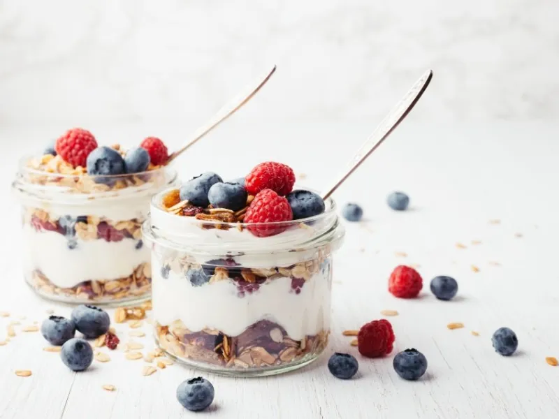 two jars with tasty parfaits made of granola, berries and yogurt on white wooden table shot at angle
