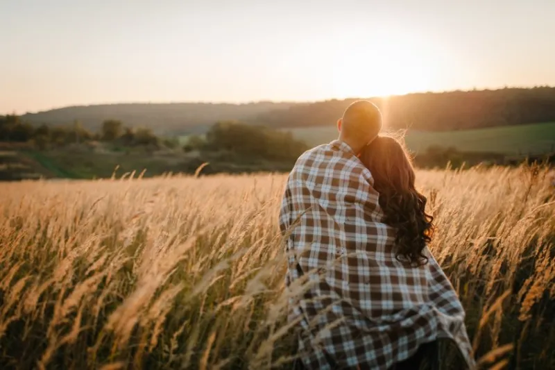 young couple hugging, standing back, people covered with blanket, at sunset in autumn an outdoor at field grass on background of sun concept of friendly family full length close up
