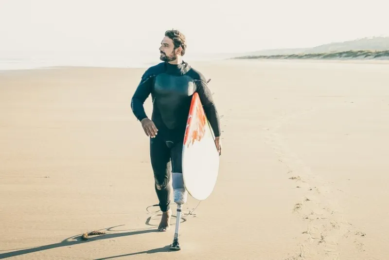 active surfer with amputated leg walking on beach with surfboard bearded amputee in wetsuit pacing on sand, carrying board and looking away vertical shot artificial limb and extreme sport concept