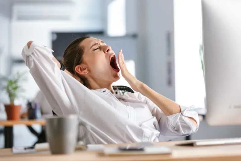 sleepy young woman dressed in shirt sitting at her workplace at the office, yawning