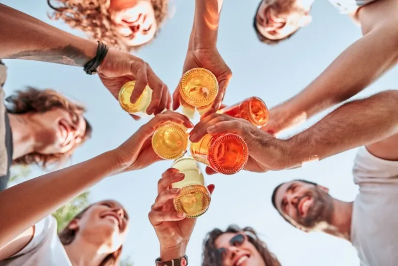 bottom view of group of happy young people clinking bottles of beer in front of blue sky