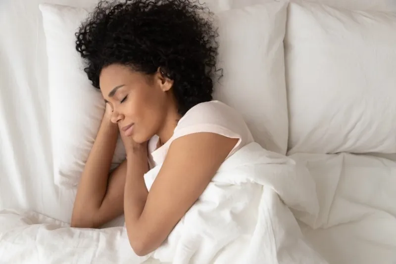 beautiful african american woman with hands under cheek sleeping in bed under blanket top view, girl with closed eyes resting in bedroom, enjoying fresh bedclothes, lying on soft pillow close up