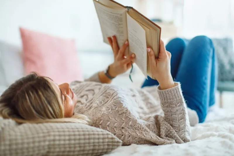 young woman lies on her back in bed reading a book