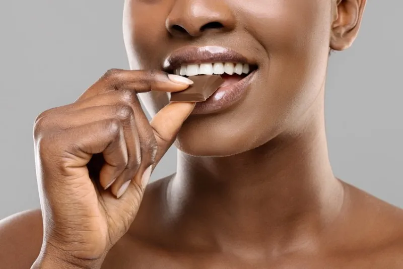 closeup portrai of black woman with white healthy teeth biting milk chocolate slice over gray background, crop
