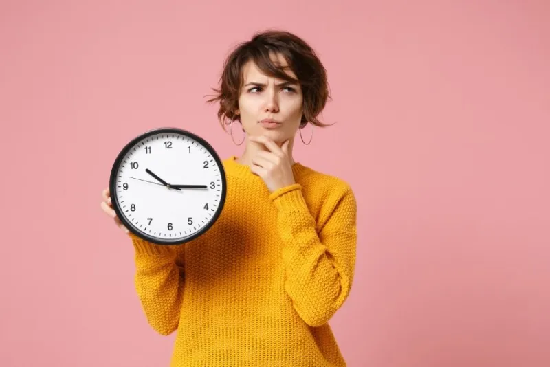 pensive young brunette woman girl in yellow sweater posing isolated on pastel pink background, studio portrait people lifestyle concept mock up copy space holding clock, put hand prop up on chin