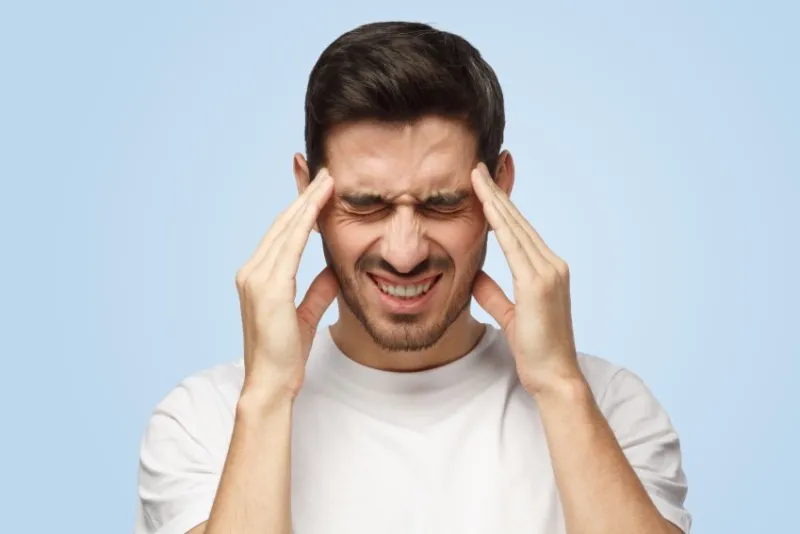 portrait of young man isolated on blue background suffering from severe headache, pressing fingers to temples, closing eyes to relieve pain with helpless face expression