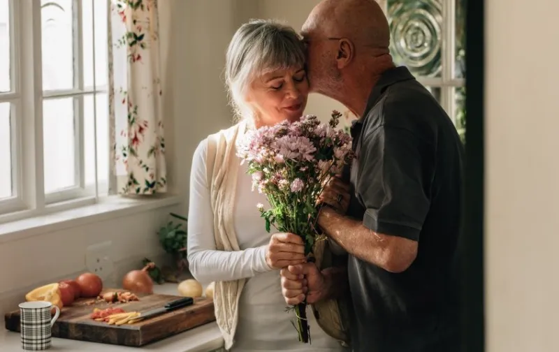 senior couple standing in kitchen holding a bunch of flowers senior man kissing his wife holding her hand at home