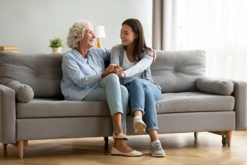 happy adult granddaughter and senior grandmother having fun enjoying talk sit on sofa in modern living room, smiling old mother hugging young grown daughter bonding chatting relaxing at home together