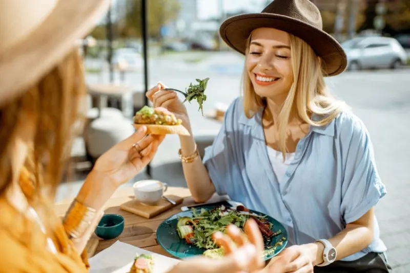 two female best friends eating healthy food while sitting together on a restaurant terrace on a summer day