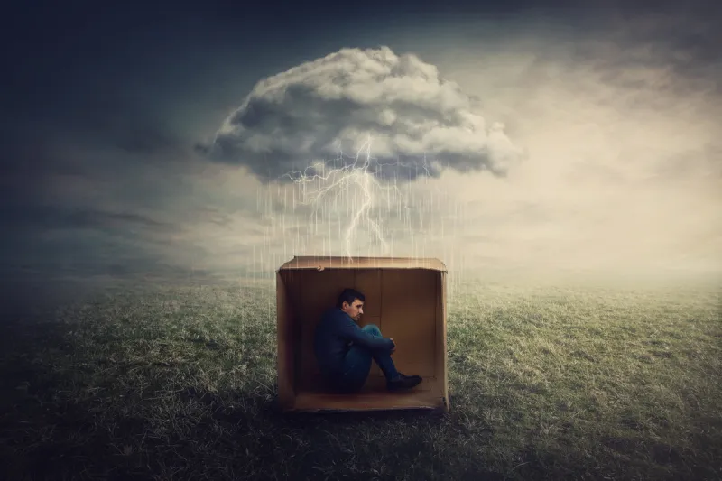 surreal concept with a scared guy shelters inside a cardboard box introvert man caged by own fears as a thunderstorm cloud trapped him under the rain mysterious storm as emotional crisis symbol