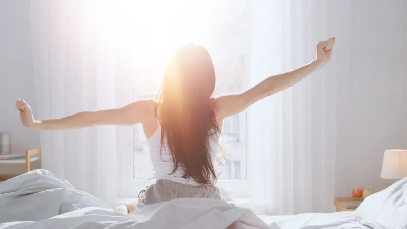 beautiful brunette is waking up in the morning, stretches in the bed, sun shines on her from the big window happy young girl greets new day with warm sunlight flare