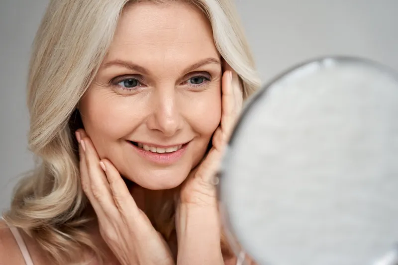 happy 50s middle aged woman model touching face skin looking in mirror smiling mature older lady pampering, enjoying healthy skin care, aging beauty, skincare treatment cosmetic products concept