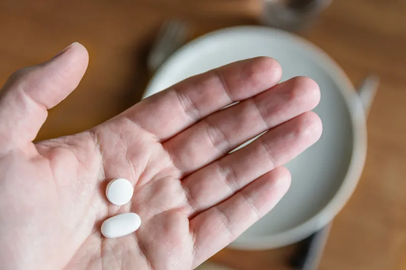 close-up view of a white male hand holding two white pills in the palm above a blurry background showing a dining table with plate, cutlery and glass