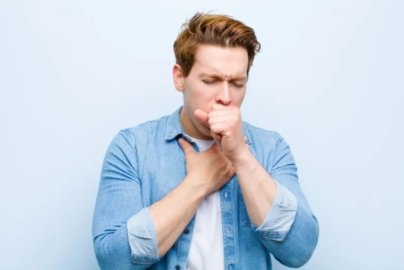 young red head man feeling ill with a sore throat and flu symptoms, coughing with mouth covered against blue wall