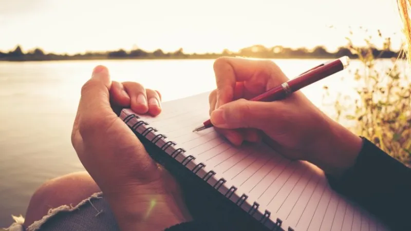 close up hand of young woman with pen writing on notebook at riverside in the evening