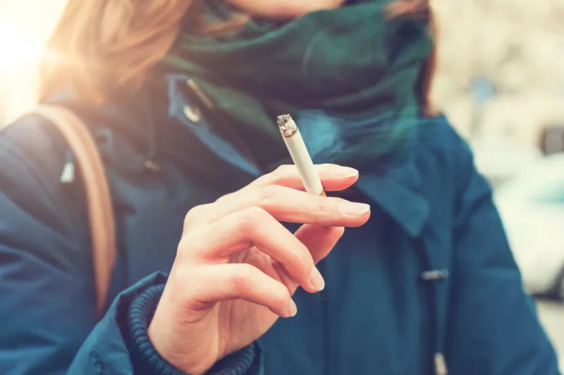 young woman enjoying a cigarette outdoors holding it between her fingers, low angle view against the chest of a warm autumn jacket in a smoking and tobacco concept