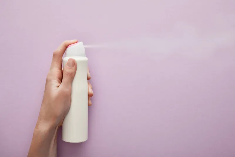 cropped view of woman spraying deodorant on violet background with white roses