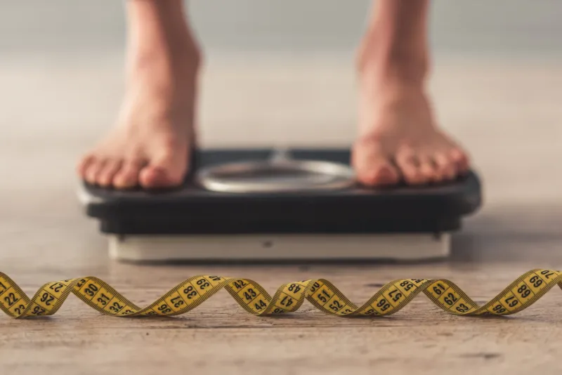 cropped image of woman feet standing on weigh scales, on gray background a tape measure in the foreground
