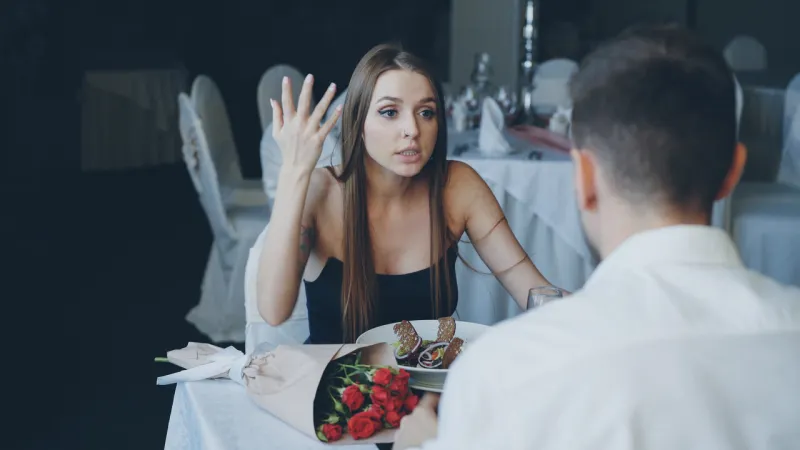 angry young woman is quarreling with her lover while dining in restaurant, shouting and gesturing emotionally bouquet of flowers and plate with food are visible
