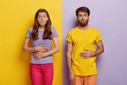 young female and male touch stomaches, feel satiety after eating hearty meal, bites lips, have good appetite, dressed in casual outfit, pose against yellow and purple wall pleasant feeling in belly