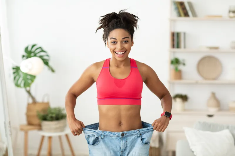 successful weight loss happy african woman after slimming showing her old oversize pants standing indoors, smiling to camera