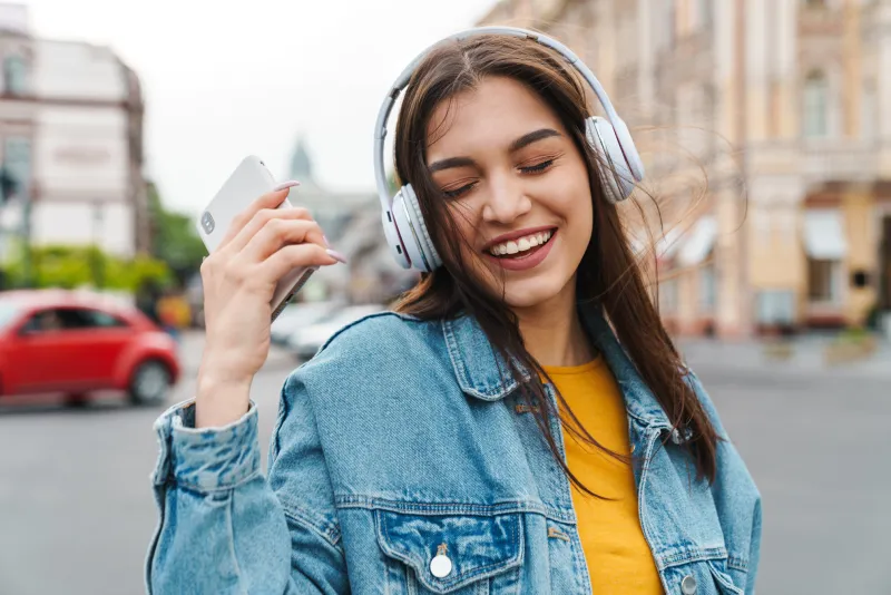 image of nice laughing woman listening music with smartphone and wireless headphones on city street