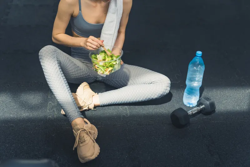 being healthy involves a diet unrecognizable caucasian young woman in gray sportswear sitting on the dark floor next to a water bottle, eating green salad and vegetables high quality photo