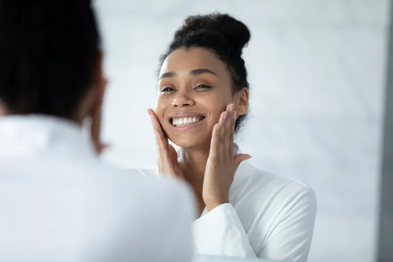 head shot happy beautiful young african american woman in white bathrobe touching face, feeling satisfied with moisturized healthy skin condition after morning spa treatment procedures in bathroom