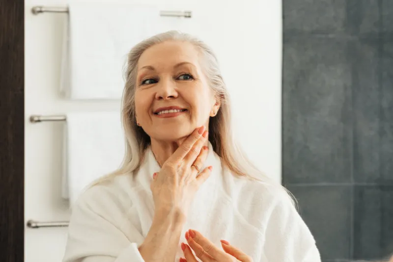 smiling senior woman touching skin on her neck in front of a mirror