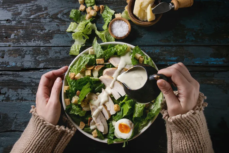female hands powring dressing to classic caesar salad with chicken breast in white ceramic plate served with ingredients above over old dark blue wooden background flat lay rustic style