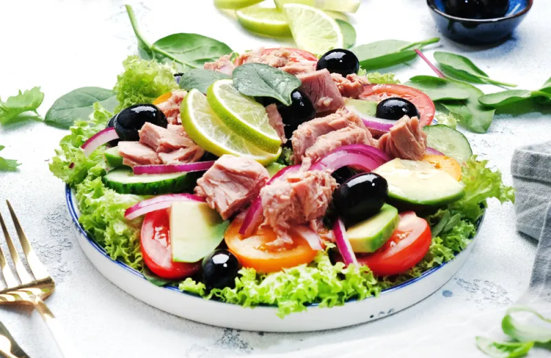 fresh tuna salad with avocado, red tomato, cucumber, black olives, spinach, onion and lettuce white table background, top view