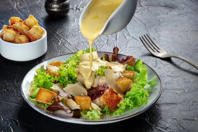 chicken caesar salad with the classic dressing being poured, croutons, and pepper, on a black background