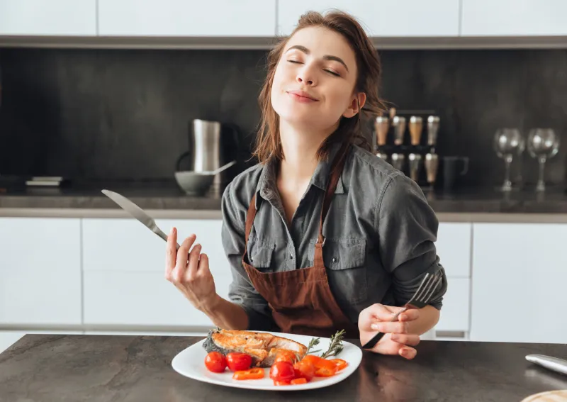 image of pretty young woman sitting in kitchen while eating fish and tomatoes