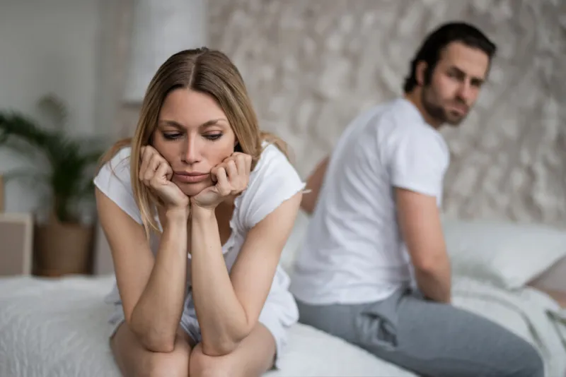 sexual problems, relationship crisis concept stressed young woman sitting on bed, overwhelmed with family difficulties, her offended husband on background, blank space
