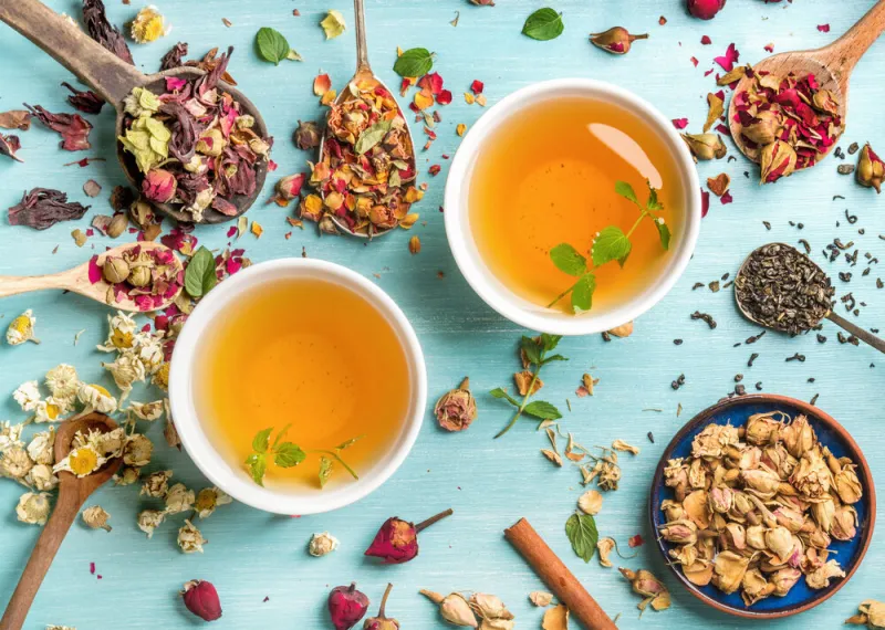 two cups of healthy herbal tea with mint, cinnamon ,dried rose and camomile flowers in spoons over blue background, top view
