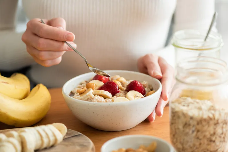 woman eating oatmeal porridge with banana, strawberries and nuts healthy breakfast at the sunny morning kitchen table