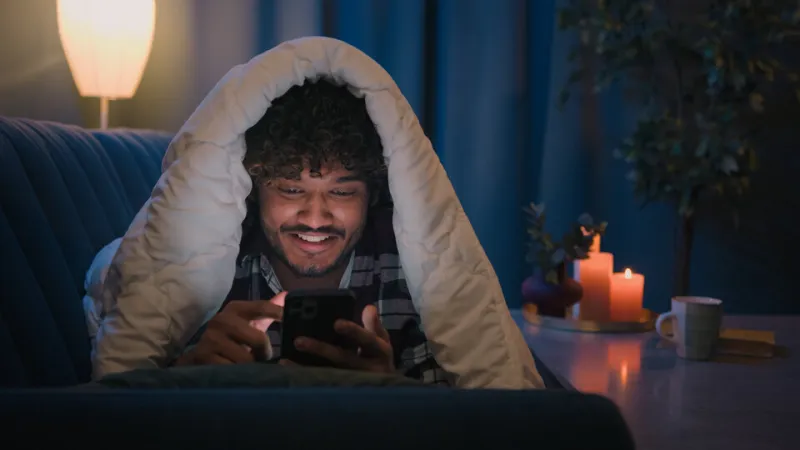 smiling guy indian man arabian male at night evening home on couch under blanket cover with duvet looking mobile phone wow surprised amazed reading news gadget addict smartphone social media scrolling high quality 4k footage