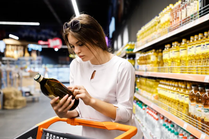 young pretty girl is shopping in a big store the girl buys groceries at the supermarket the girl chooses oil in a glass bottle