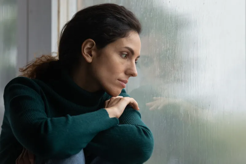 thoughtful stressed young hispanic latin woman sitting on windowsill, looking outside on rainy weather, having depressive or melancholic mood, suffering from negative thoughts alone at home