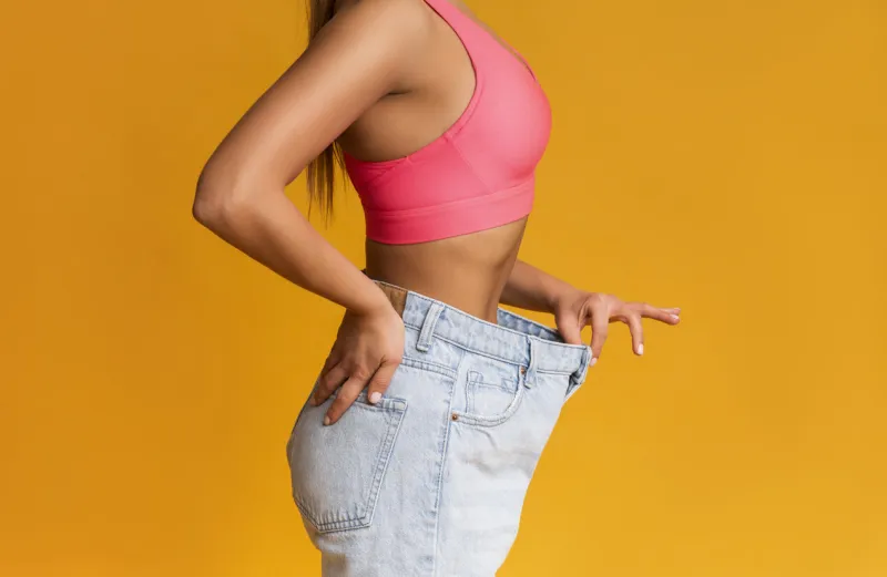 weight loss concept profile portrait of fit girl posing in oversize jeans over yellow background, cropped image with copy space