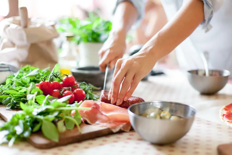 woman is cooking in home kitchen female hands cut salami, vegetables, greens, tomatoes on table on wooden boards ingredients for preparing italian or french food lifestyle moment