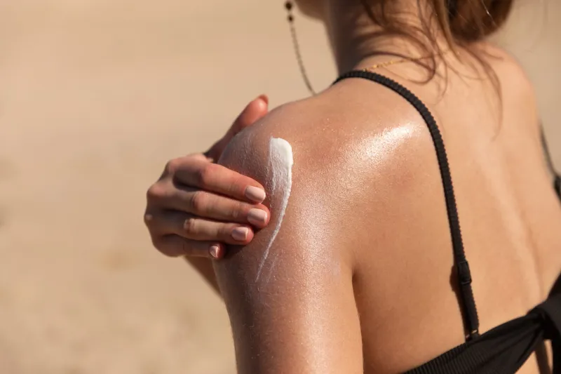 a young woman applying sun cream or sunscreen on her tanned shoulder to protect her skin from the sun shot on a sunny day with blurry sand in the background