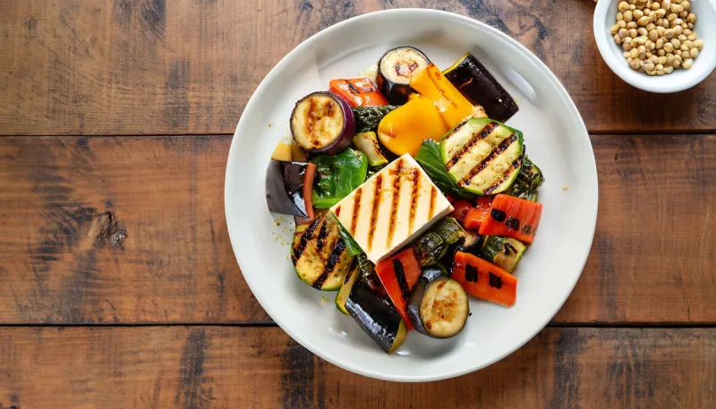 overhead view of a plate of healthy grilled roast vegetables with tofu, or soybean curd, on a wooden table