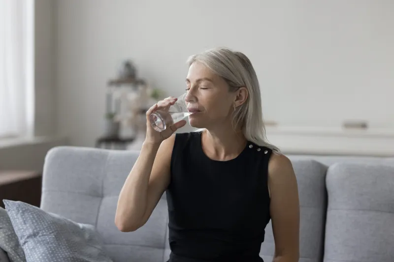 peaceful pretty blonde mature woman drinking natural mineral water with closed eyes for healthcare, healthy metabolism, refreshment, sitting on couch at home, satisfying thirst