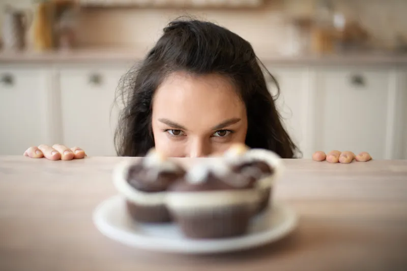 young woman hiding behind table and looking at delicious cupcakes on plate, focus on lady appetite, diet and gluttony concept