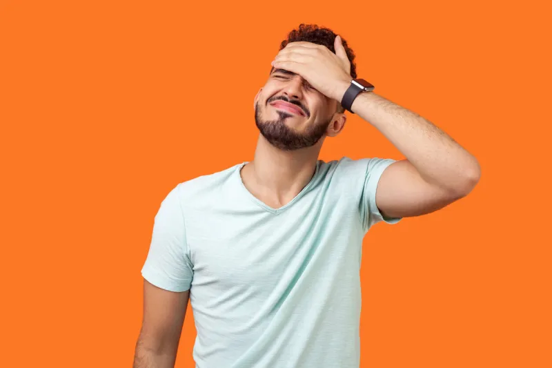 facepalm portrait of desperate brunette man with beard in white t-shirt covering face with hand, feeling sorry and blaming himself for the mistake indoor studio shot isolated on orange background