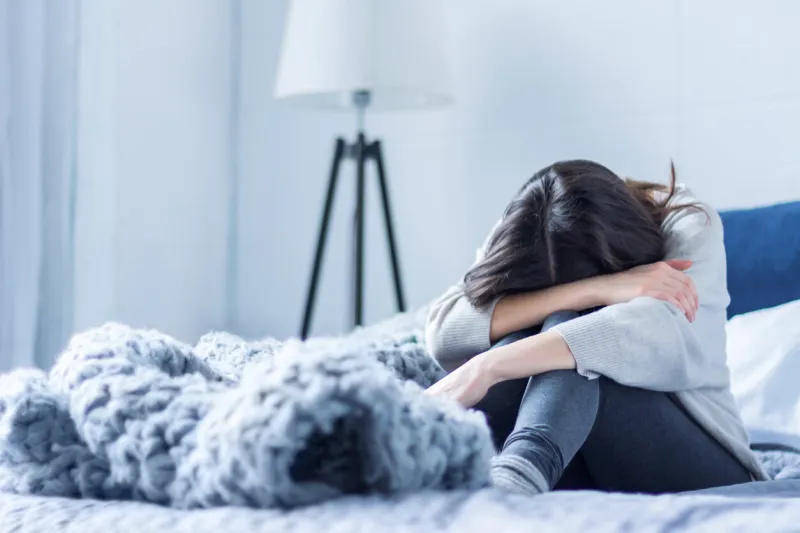 young woman in gray clothes is sitting curled up on bed at home upset brunette girl is suffering, crying depression because of loneliness, stress, problems psychological disorders concept